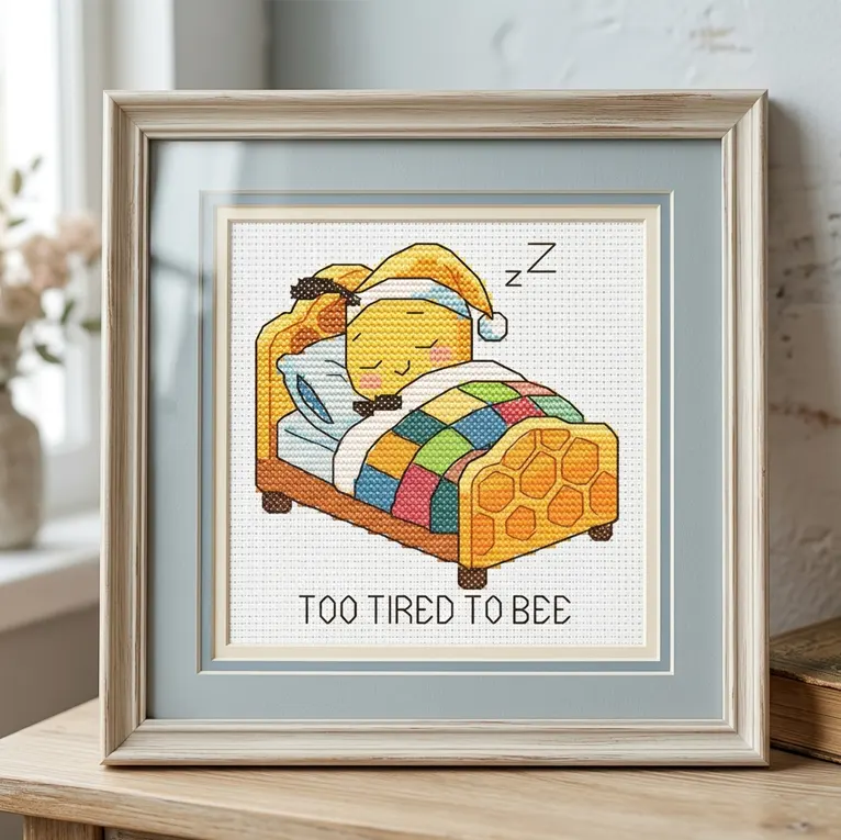 Framed cross stitch of a sleeping bee in a honeycomb bed under a quilt with text on white Aida.